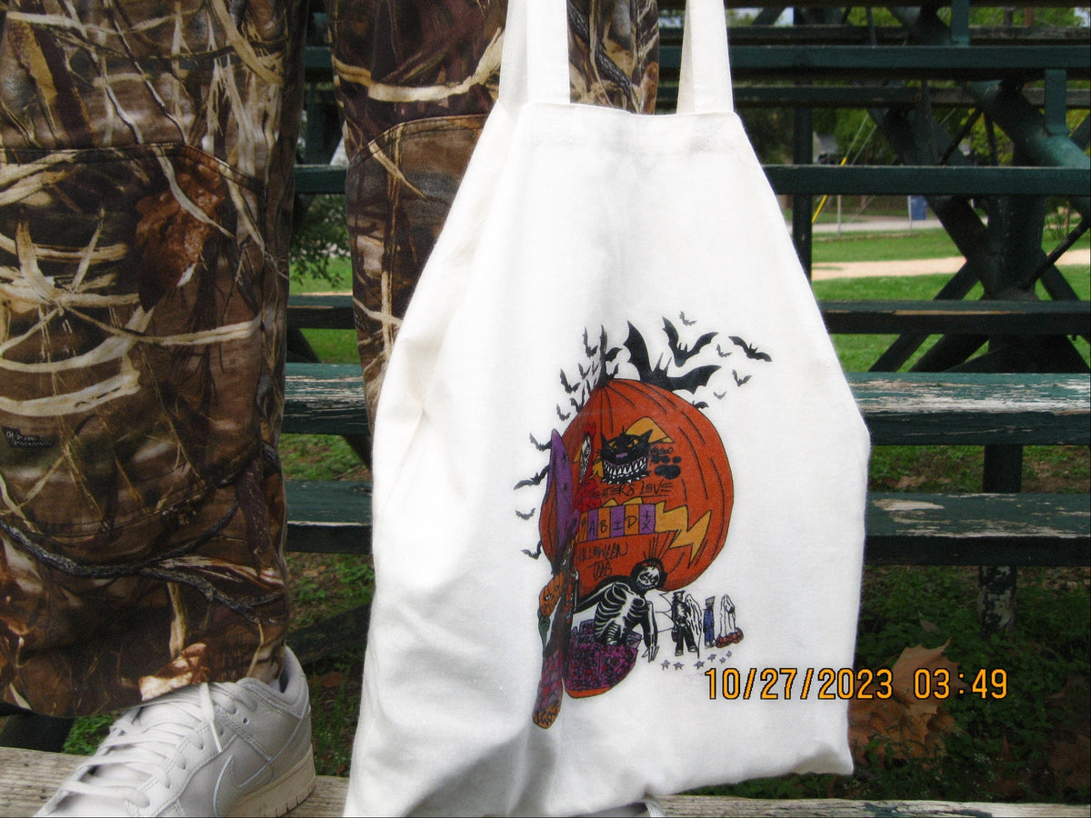 White tote bag with Halloween design held by a person wearing camouflage pants and white sneakers.