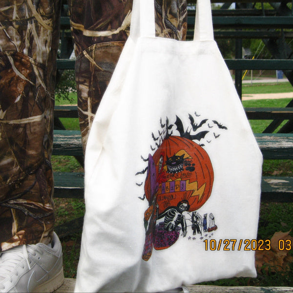 White tote bag with Halloween design held by a person wearing camouflage pants and white sneakers.