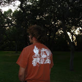 Person wearing an orange t-shirt with a graphic design, standing in a forested area.