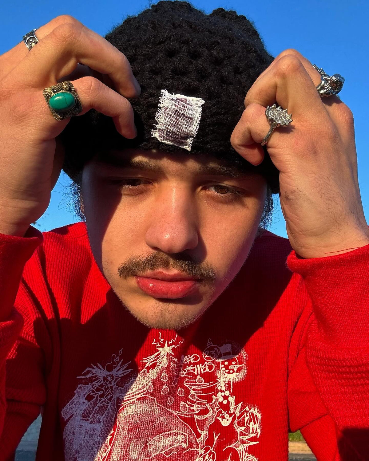 Person wearing a black knit cap with a white label, red sweater, and multiple rings against a blue sky.