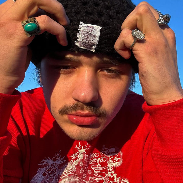Person wearing a black knit cap with a white label, red sweater, and multiple rings against a blue sky.